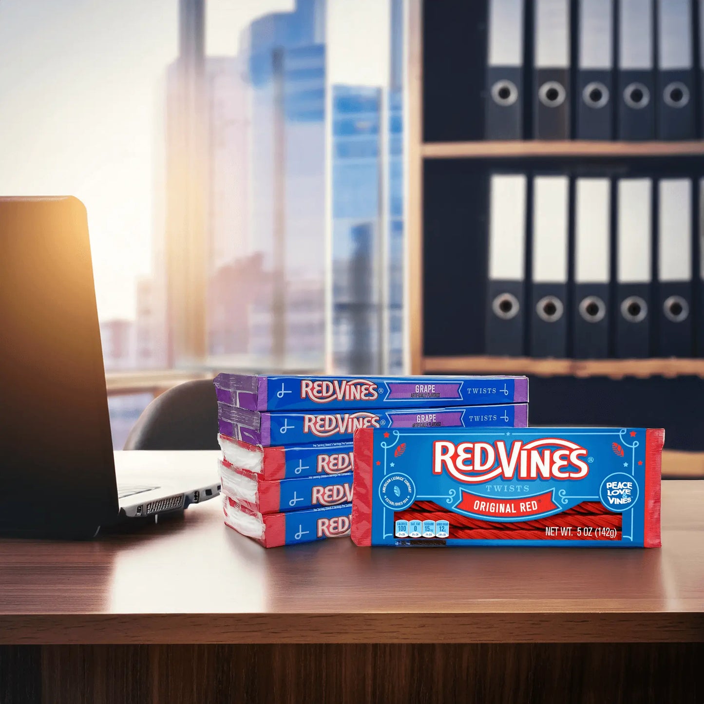 A stack of Red Vines trays on an office desk with a city view of the sun setting through the window