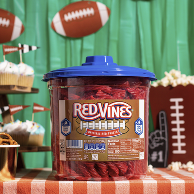 A large plastic jar of Red Vines Original Red Twists candy with football label and blue lid, set on a checkered tablecloth with football-themed party decorations in the background.