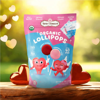 Torie & Howard Organic Valentine's Candy Lollipops on wooden table with sunshine and illuminated hearts in the background