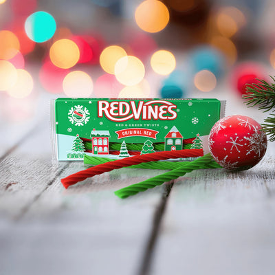 Red Vines Twists packaging with a Christmas-themed design on a wooden surface with red and green colored licorice candy in front and blurred lights in the background.