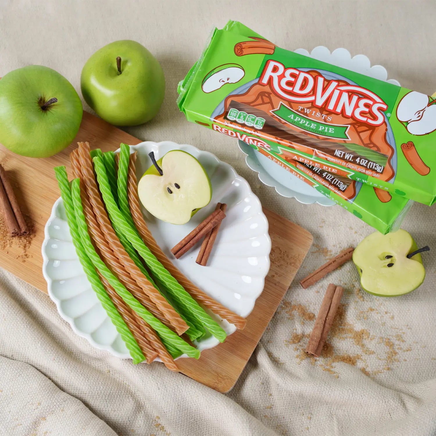 Red Vines Apple Pie flavored twisted candy sticks displayed on a white scalloped plate with green apples, cinnamon sticks, and the green product packaging on a beige fabric background.
