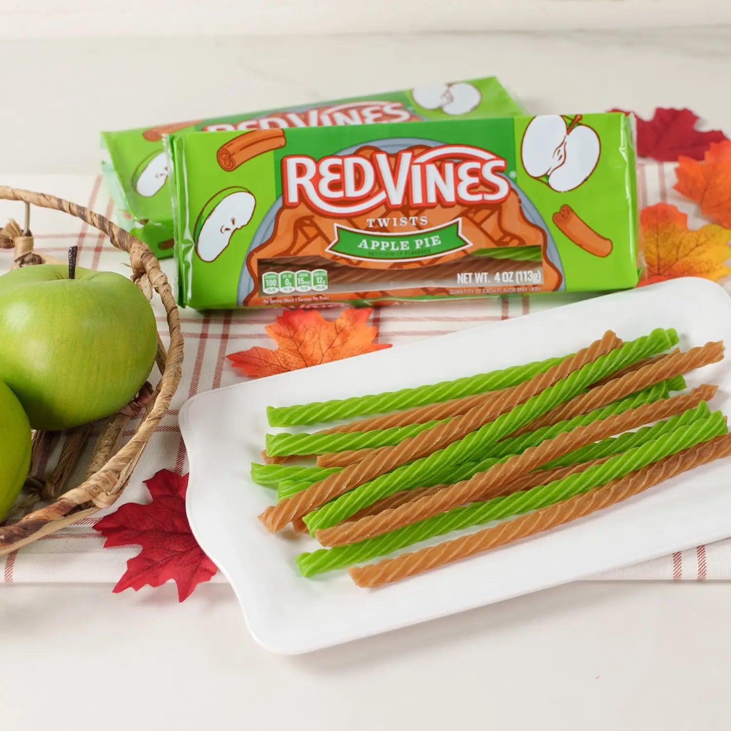 Red Vines Apple Pie Twists candy sticks on a white plate, with two green packages of the candy behind them and green apples and autumn leaves on a white surface.