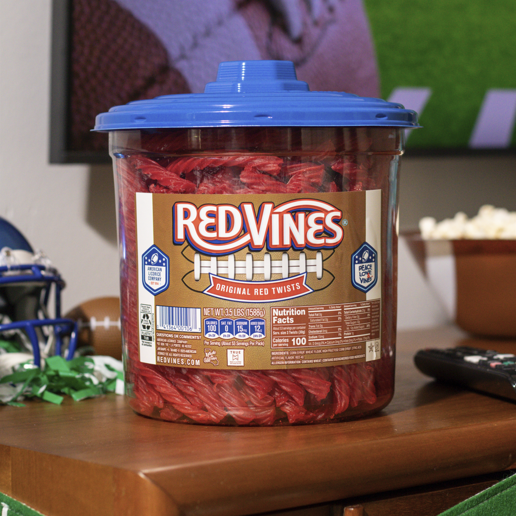 A large plastic tub of Red Vines Original Red Licorice candy with a blue lid and football label, sitting on a wooden table in a living room setting with a remote control, popcorn bowl, and football game on tv in the background.