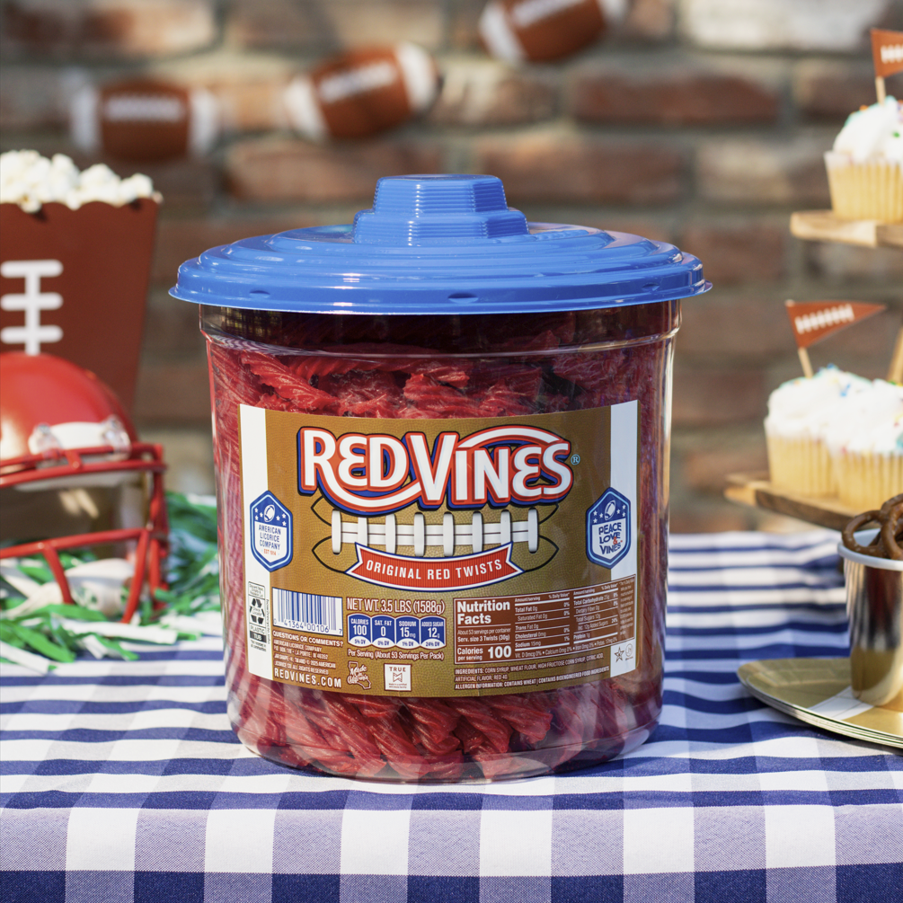 A large jar of Red Vines Original Red Licorice Twists candy with a football label and blue lid, set on a blue-and-white checkered tablecloth with football-themed party decorations in the background.