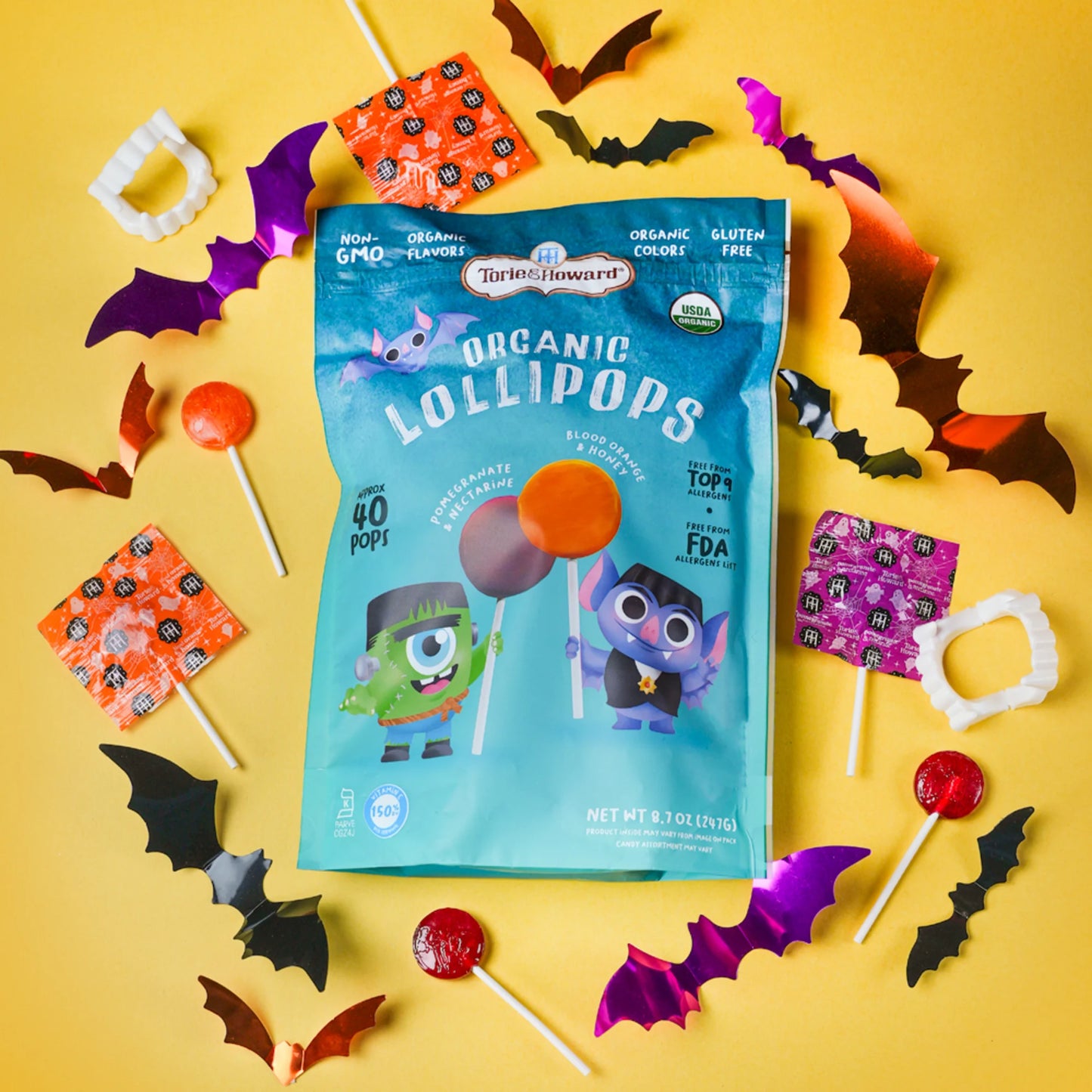 A bag of Torie & Howard Organic Lollipops is displayed in a flat-lay arrangement on a bright yellow background, surrounded by Halloween-themed decorations including bat cutouts, candy corn, and lollipops.