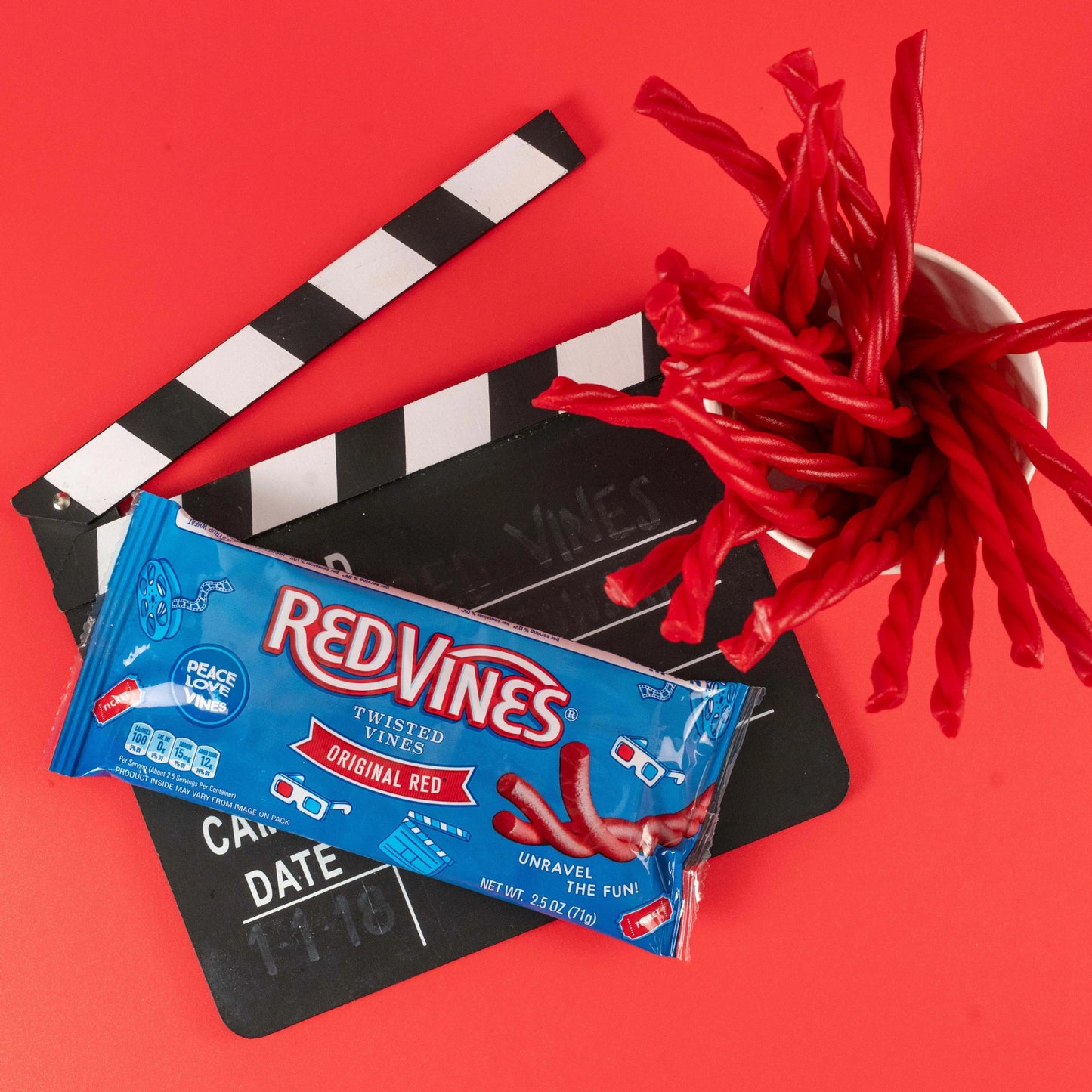 Red Vines Original Red Twisted Vines 2.5oz Bag on a red background with film clapper and raw pull and peel licorice pieces