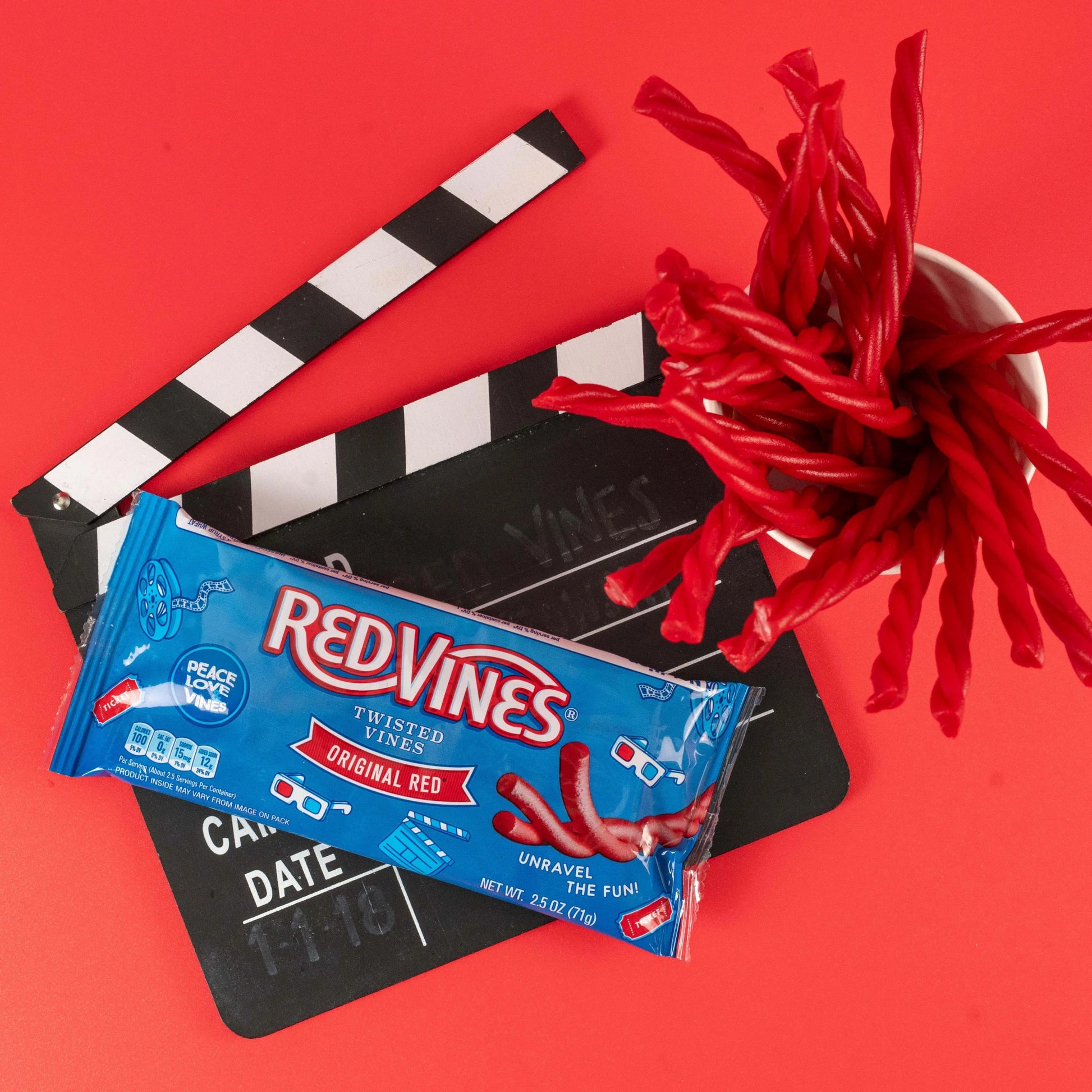 Red Vines Original Red Twisted Vines 2.5oz Bag on a red background with film clapper and raw pull and peel licorice pieces