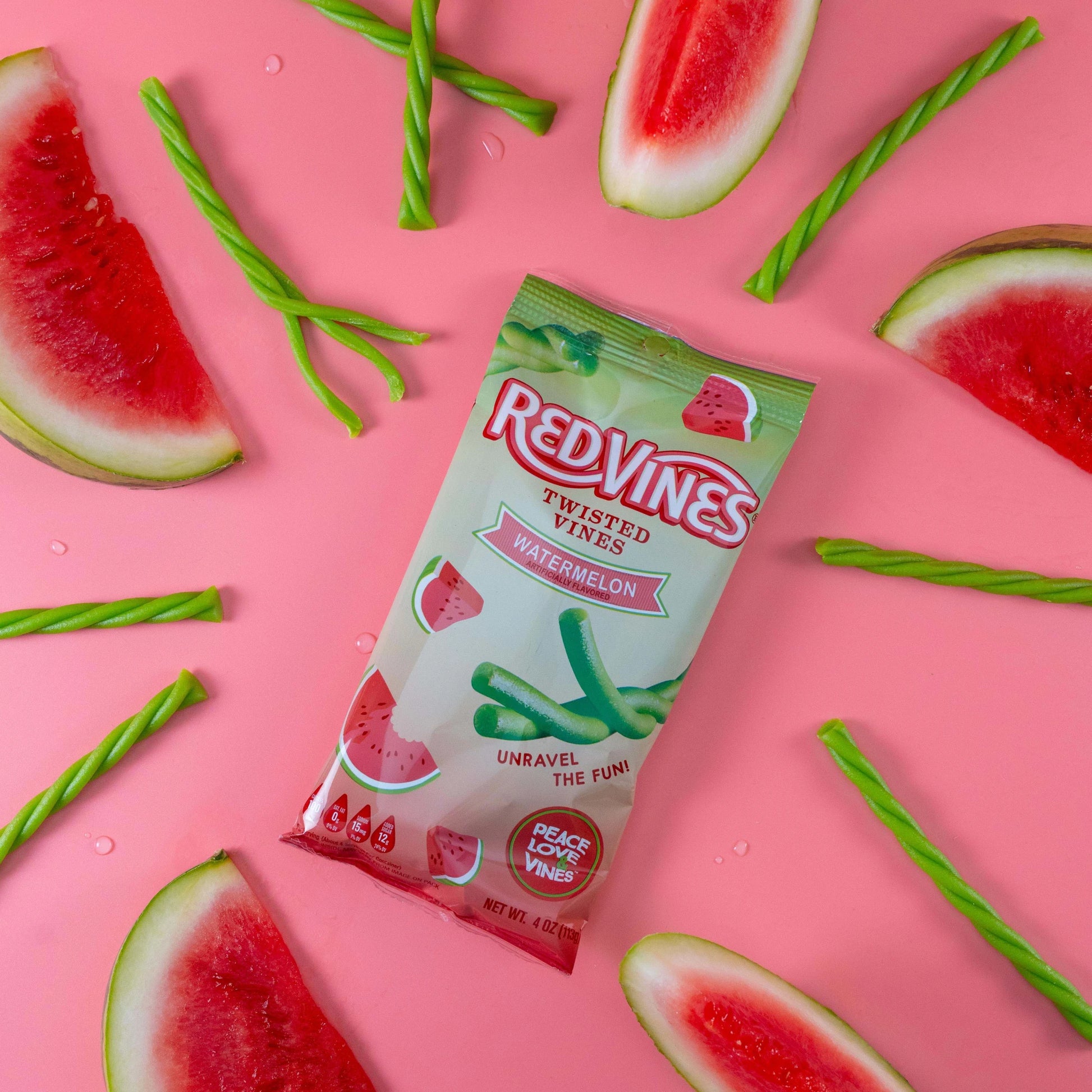 Red Vines Watermelon Twisted Vines 4oz Bag on a pink background with raw watermelon licorice pieces and real watermelon fruit