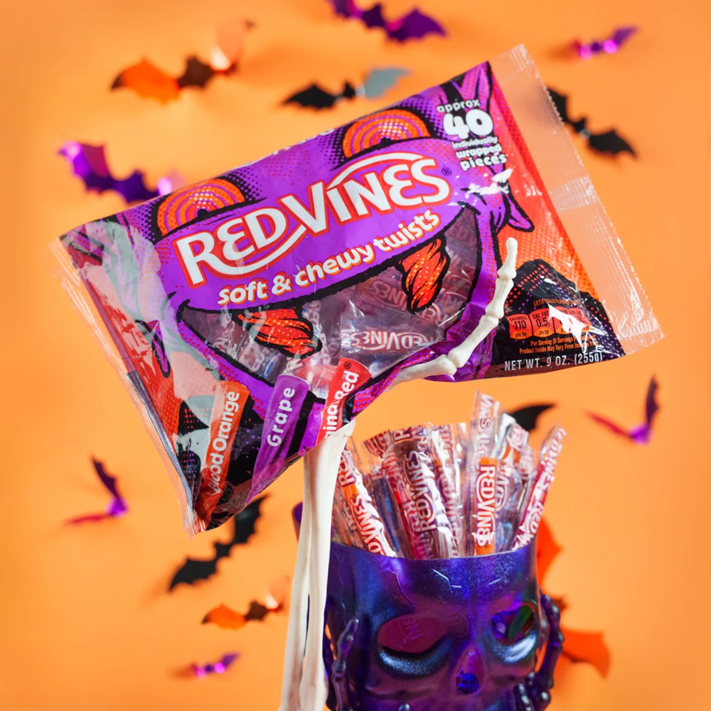 A bag of Red Vines Soft & Chewy Twists candy is shown floating above a purple skull-shaped container filled with assorted Red Vines lollipops, set against an orange background with scattered Halloween bat decorations.