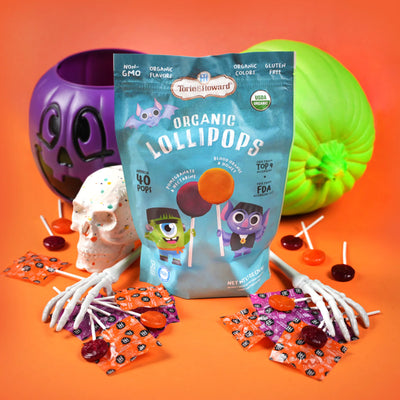 A bag of Torie & Howard Organic Lollipops displayed on an orange surface with Halloween props, including a purple pumpkin, a green pumpkin, a skull, skeleton hands, and scattered wrapped lollipops.