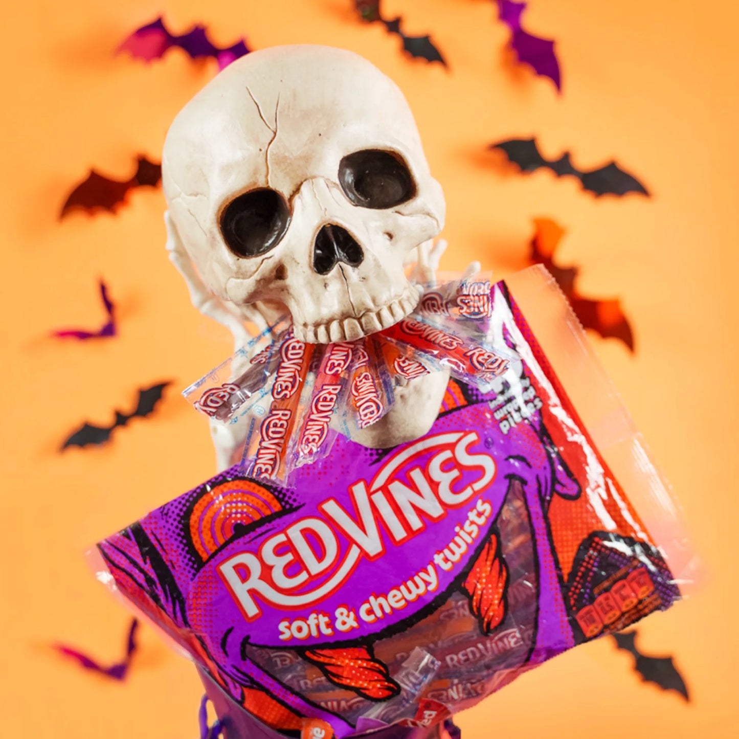 A Halloween-themed product shot featuring a plastic skull holding a bag of Red Vines Soft & Chewy Twists candy against an orange background with black bat cutouts.